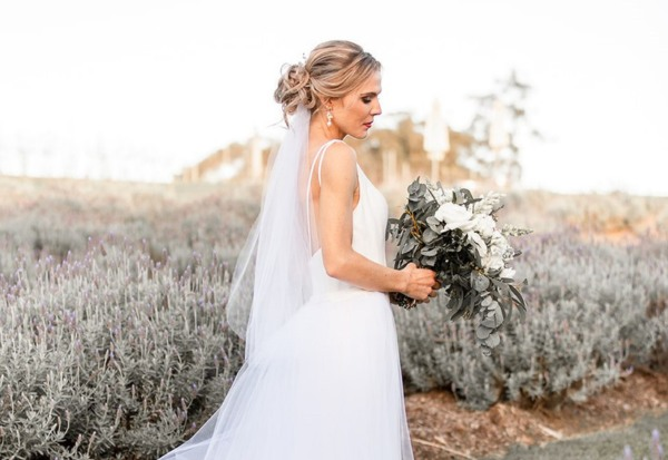 Blonde woman with styled bridal messy bun and glam makeup in wedding dress and veil, standing with floral arrangement in lavender field.