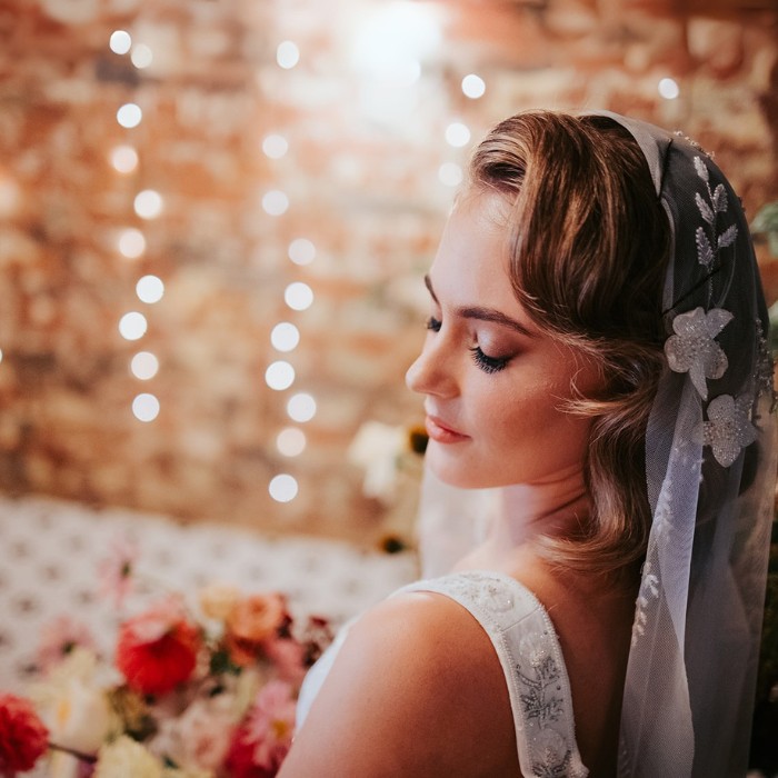 Glam makeup with dark whispy lashes and soft wavy hair syled with floral beaded veil and fairy lights nighttime background.