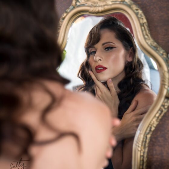 Glam makeup with winged liner and red lips on brunette woman with styled long wavy curls looking at herself in gilded gold mirror. Art deco aesthetic.