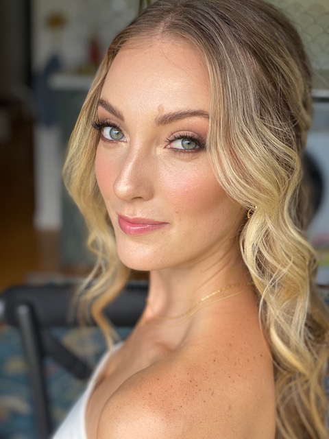 Soft smiling blonde woman with styled long wavy curls, soft glam makeup with rosy nude lipstick.