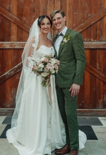 Smiling young wedding couple, woman with dark hair and long white veil and dress, man with moustashe and blonde hair, plaid green and grey suit standing in front of barn door.