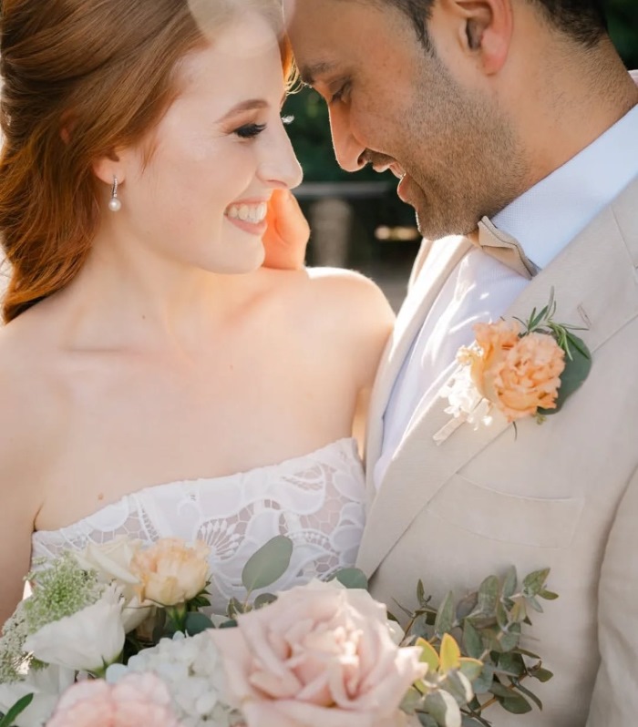 Smiling woman with auburn red hair and man with 1 day shave growth and dark hair. Wedding styling with pastel peach and pink floral bouquet.