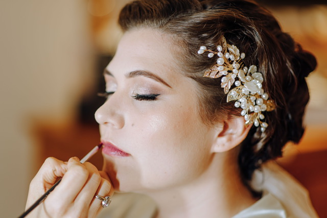 Woman with closed eyes upstyled dark hair in bun and beaded gold hairpiece, having pink lipstick applied with lip brush.